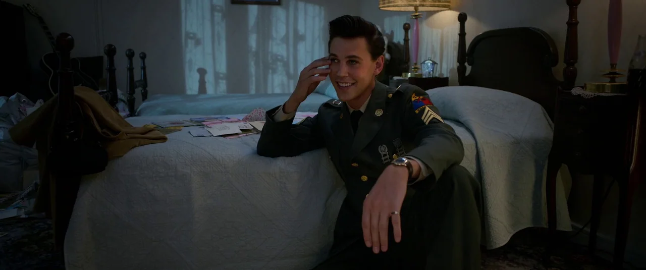 Elvis (2022) movie still: medium — A dark bedroom features a quilted bed covered with several pieces of mail. A young man in a military uniform…