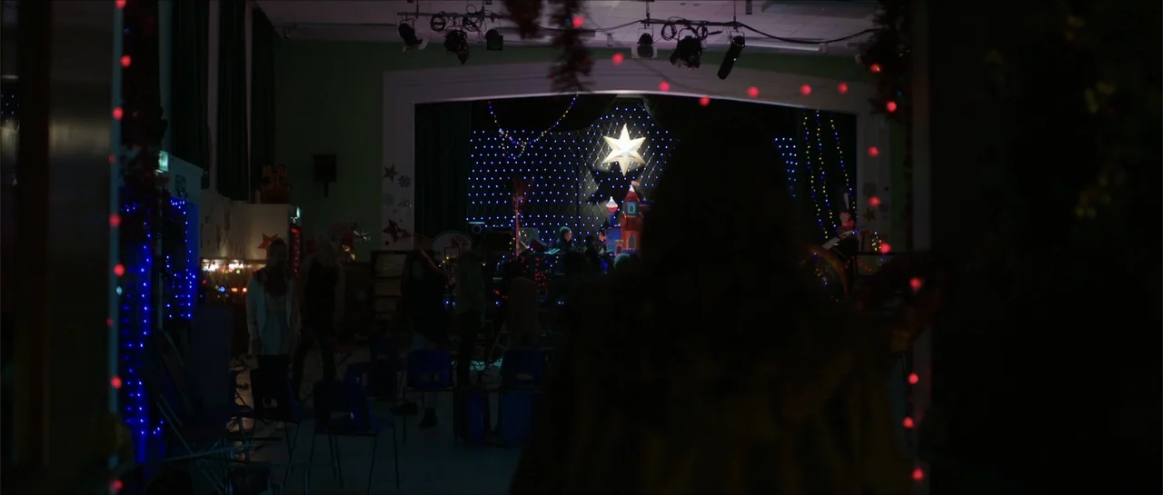Anna and the Apocalypse (2018) movie still: medium — A darkened room decorated with Christmas lights shows people gathered on a distant stage. A…