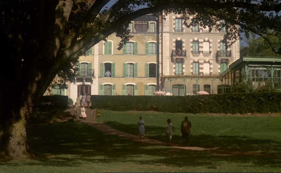 Murmur of the Heart (1971) movie still: wide — A large stone manor house with green shutters sits behind a manicured hedge. Several people walk on a…