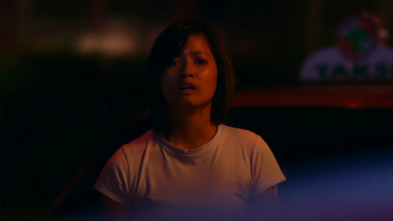 Merantau (2009) movie still: medium — A woman stands on a city street at night under warm artificial lighting. A taxi sign is visible in the blurred…