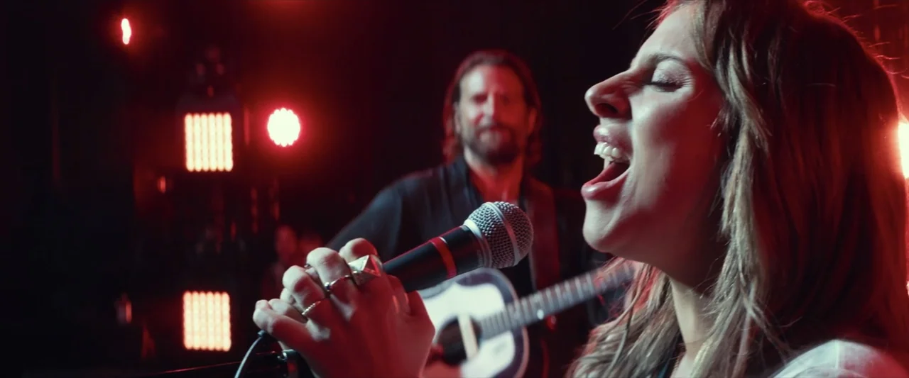 A Star Is Born (2018) movie still: closeup — A dark concert stage is illuminated by rows of bright red stage lights. A woman sings into a handheld…