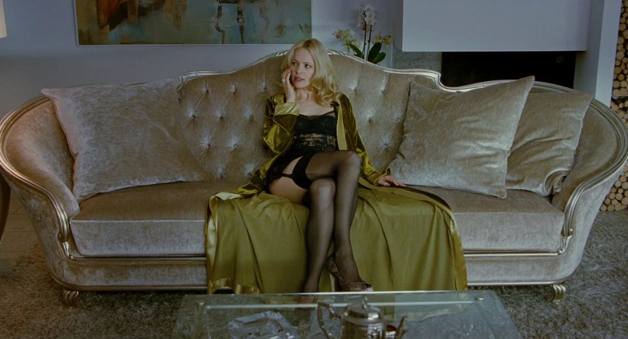 Passion (2012) movie still: medium — A modern, upscale space features a tufted velvet sofa against a neutral wall. A blonde woman in a silk robe…