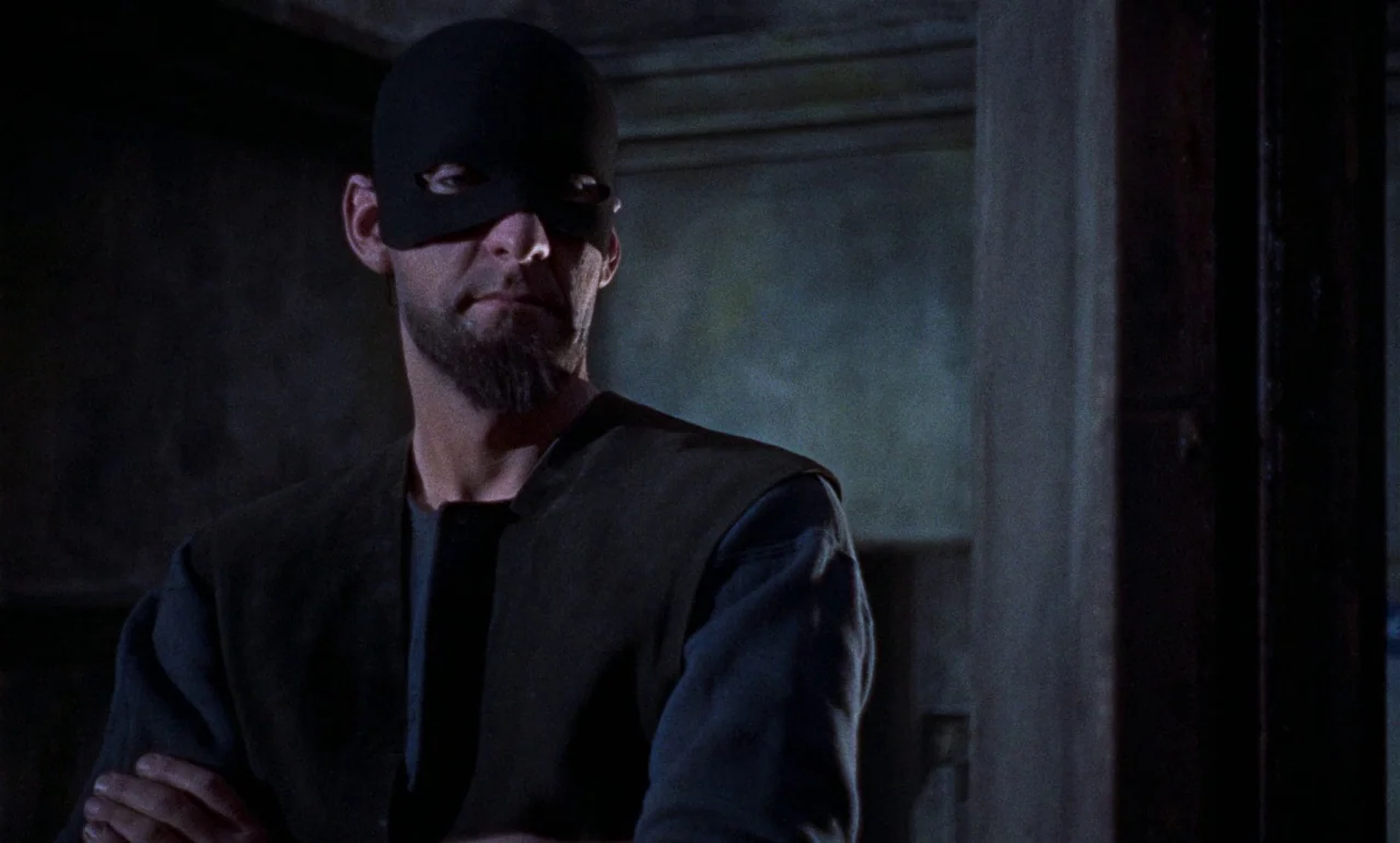 The Revenge of Frankenstein movie still: medium — A dimly lit indoor space with textured walls and deep shadows. A man wearing a black mask stands…