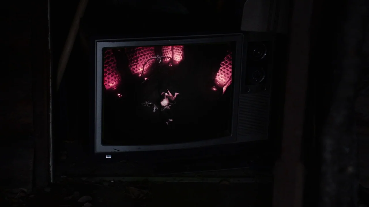 Snowbound (2017) movie still: closeup — A dark, dimly lit room contains an old CRT television set sitting in shadows. The television screen emits a…