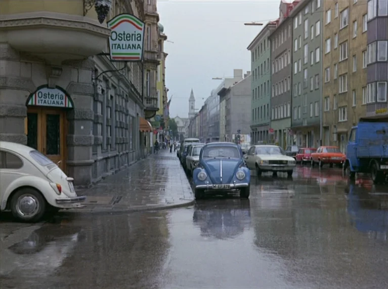 Ali: Fear Eats The Soul (1974) movie still: establishing — An overcast European city street features wet pavement and several parked classic…