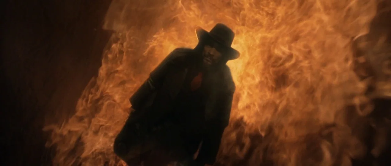 Bones movie still: medium — A man stands in a desolate area completely engulfed by a wall of fire. The intense flames illuminate his dark clothing…