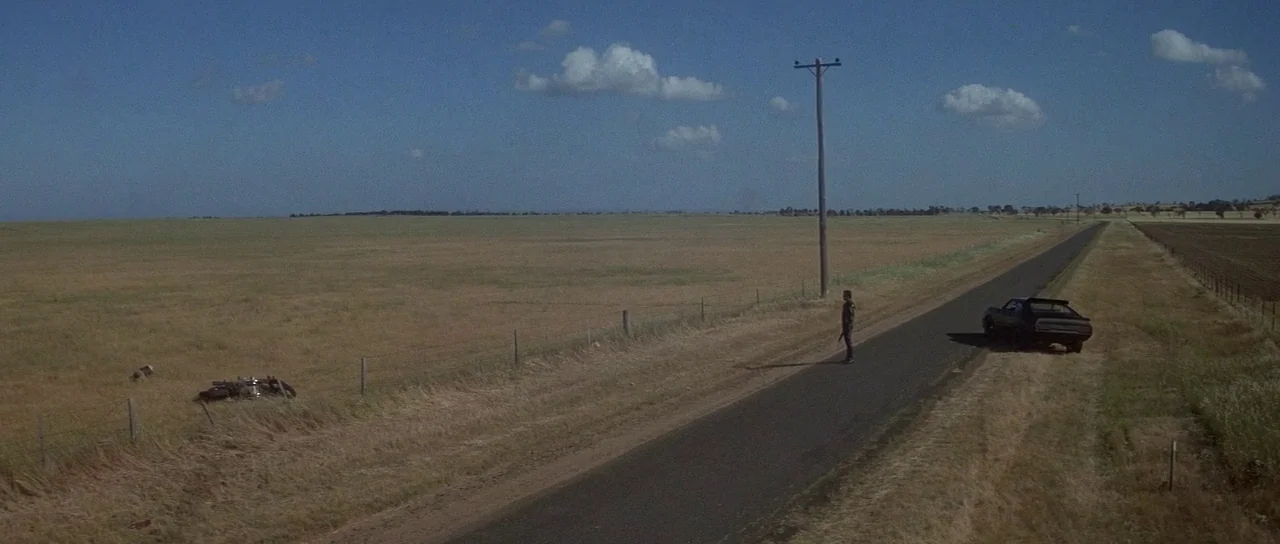 Mad Max (1979) movie still: wide — A vast, flat, grassy landscape extends toward the horizon under a clear blue sky. A single man stands on a paved…
