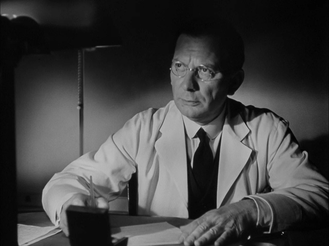 A Tree Grows In Brooklyn (1945) movie still: medium — A dimly lit room features a man sitting at a desk with paperwork. The primary light source…