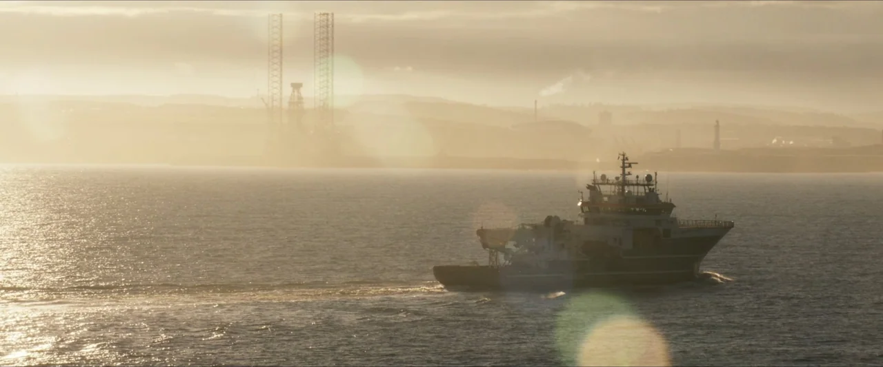 Last Breath (2025) movie still: wide — A support vessel navigates calm ocean waters during a hazy golden hour sunset. Industrial oil rig structures…