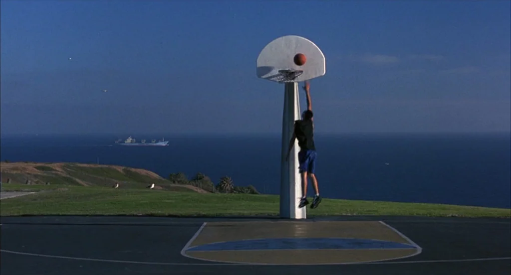 He Got Game (1998) movie still: wide — An outdoor basketball court overlooks a calm blue ocean under a clear sky. A lone player jumps to make a shot…