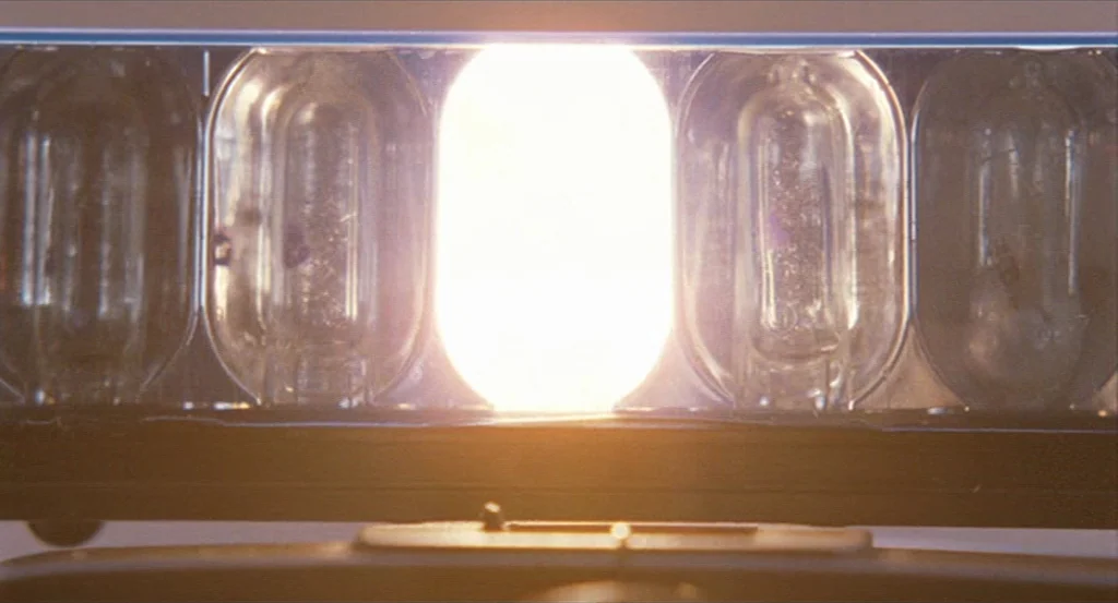 Submarine movie still: closeup — A close-up view shows the illuminated center bulb of a vehicle emergency lightbar. Glass reflectors surround the…