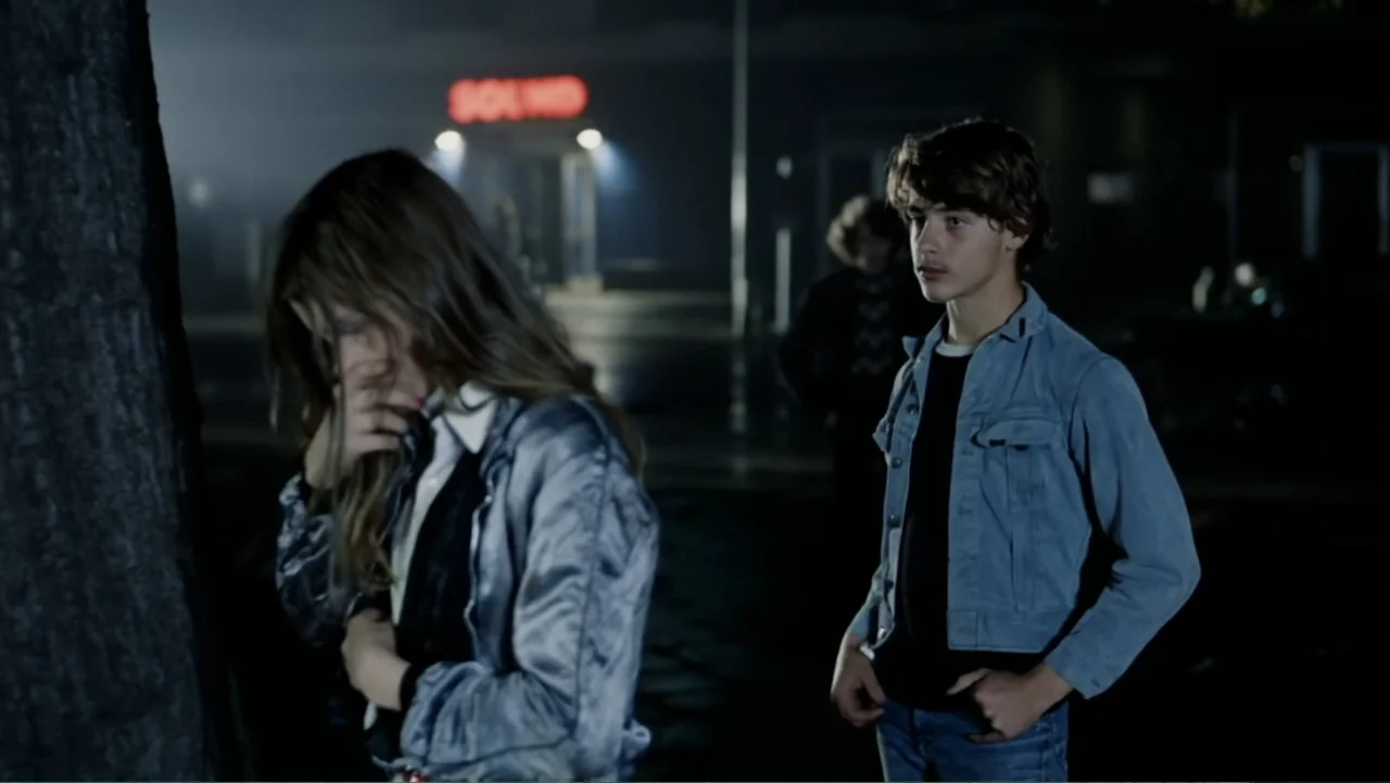 Christiane F. (1981) movie still: medium — A dark urban parking lot at night features a red neon sign in the background. A young woman stands by a…