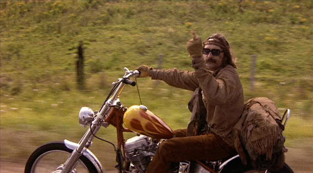 Easy Rider (1969) movie still: fullbody — A man rides a customized chopper motorcycle along a grassy rural roadside. He wears a leather fringe…