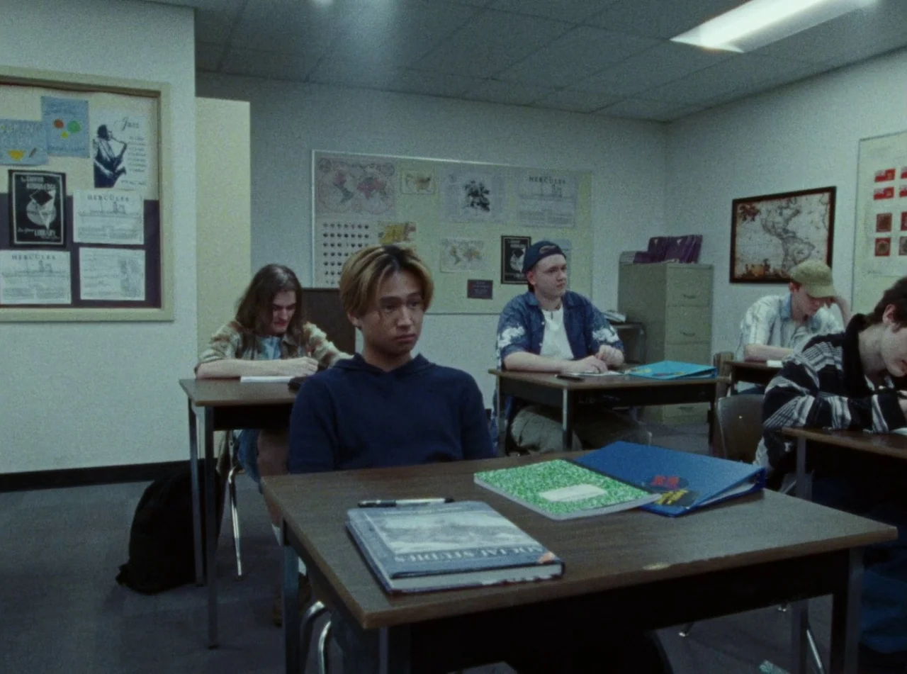 Riceboy Sleeps (2022) movie still: medium — A high school classroom features white walls decorated with maps and student posters. Several students…