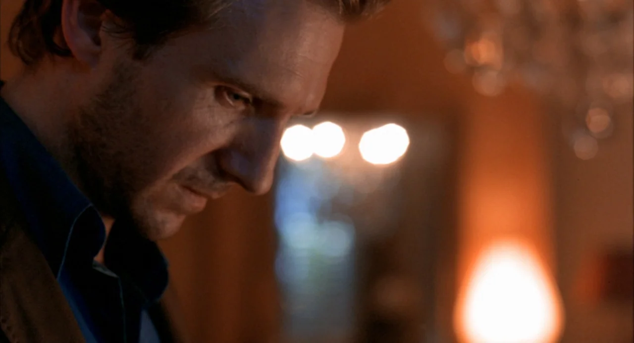 The Good Thief (2002) movie still: closeup — A man stands in a dimly lit room with blurred warm lights in background. The man looks down while…