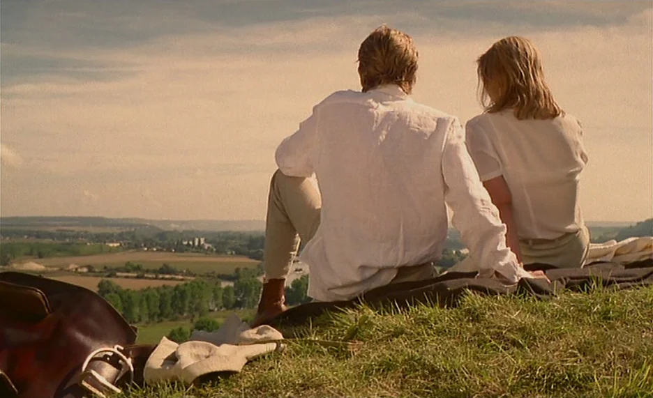 Pola X (1999) movie still: fullbody — A grassy hill overlooks a sprawling rural valley under a warm late afternoon sky. A man and a woman sit…