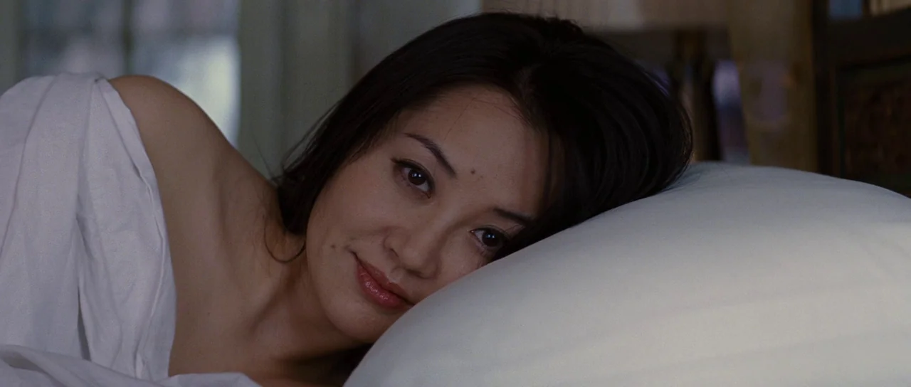 Looper (2012) movie still: closeup — A woman rests on a plush white pillow under white bed linens indoors. She gazes toward the viewer with a slight…