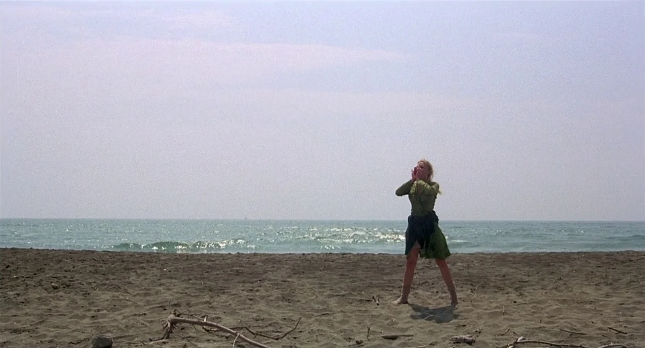 Amarcord (1973) movie still: wide — A wide beach features dark, textured sand leading to a calm sea. A lone woman stands in the center of the frame…