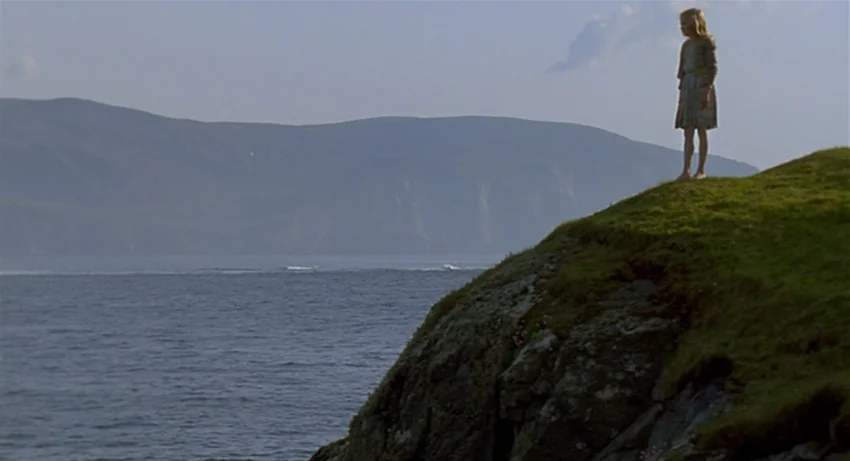 The Secret Of Roan Inish (1994) movie still: wide — A grassy cliff edge overlooks a large body of water beneath a clear sky. A young girl in a plain…
