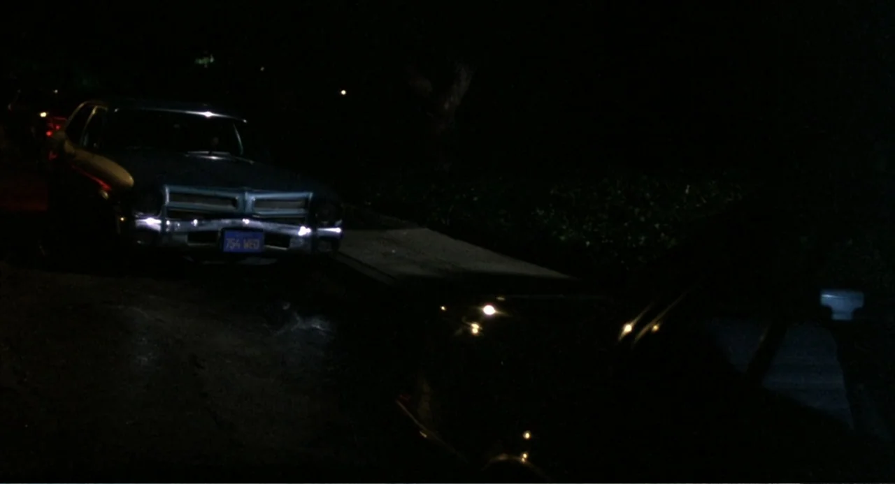 Murphy's Law (2009) movie still: wide — A dark, nighttime residential street features a parked vintage sedan in the foreground. The car's front end…