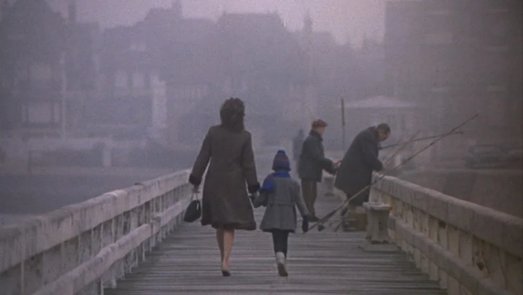 Un Homme et Une Femme (1966) movie still: fullbody — A wooden pier extends over water into a thick bank of coastal fog. A woman holding hands with a…
