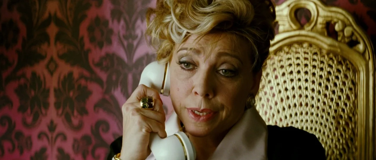 Broken Embraces (2009) movie still: closeup — A woman sits in a carved gold chair against intricate patterned crimson wallpaper. She holds a white…