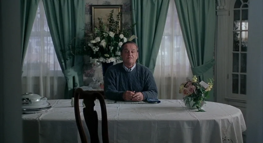 About Schmidt movie still: medium — A formal dining room features floral wallpaper and green curtains covering two windows. An older man sits at the…