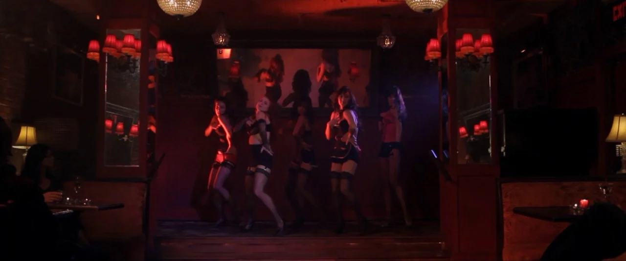 Girl Walks Into a Bar (2011) movie still: wide — A dimly lit club features a wooden stage with five women performing a dance. Red walls and antique…