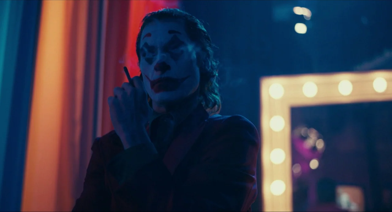 Joker (2019) movie still: medium — A dimly lit room contains a vanity mirror surrounded by bright circular light bulbs. A man in clown makeup smokes…