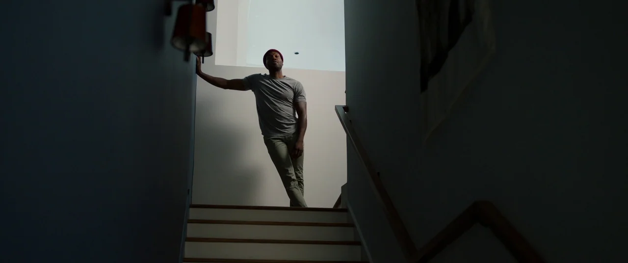 Candyman (2021) movie still: fullbody — A man stands on a residential staircase within a dark, modern house interior. He leans his body against a…