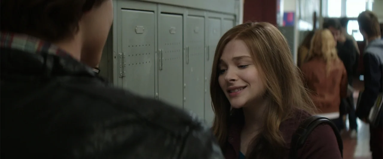 If I Stay (2014) movie still: medium — A school hallway lined with rows of grey metal lockers and students walking past. A young woman smiles while…