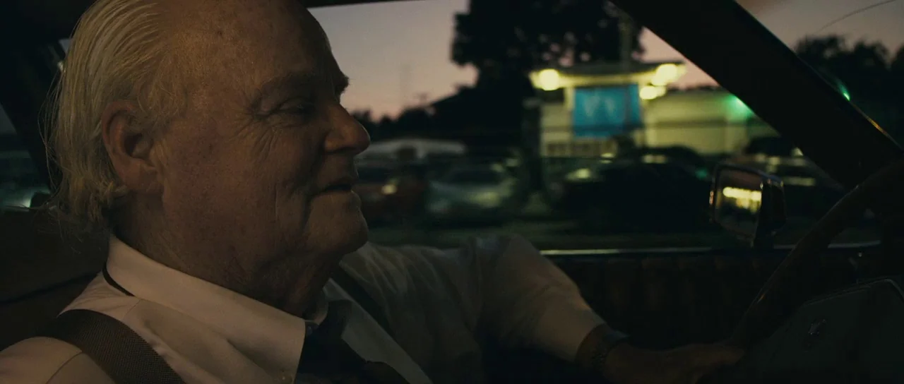 The Imposter (2012) movie still: closeup — The interior of a moving car is dark during the transition into night. An elderly man sits behind the…