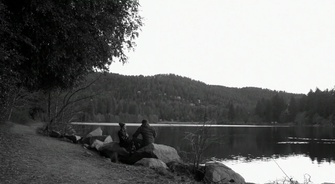 Blue Jay (2016) movie still: wide — A man and a woman sit on large boulders at the edge of a lake. A dense forest covers the mountains in the…