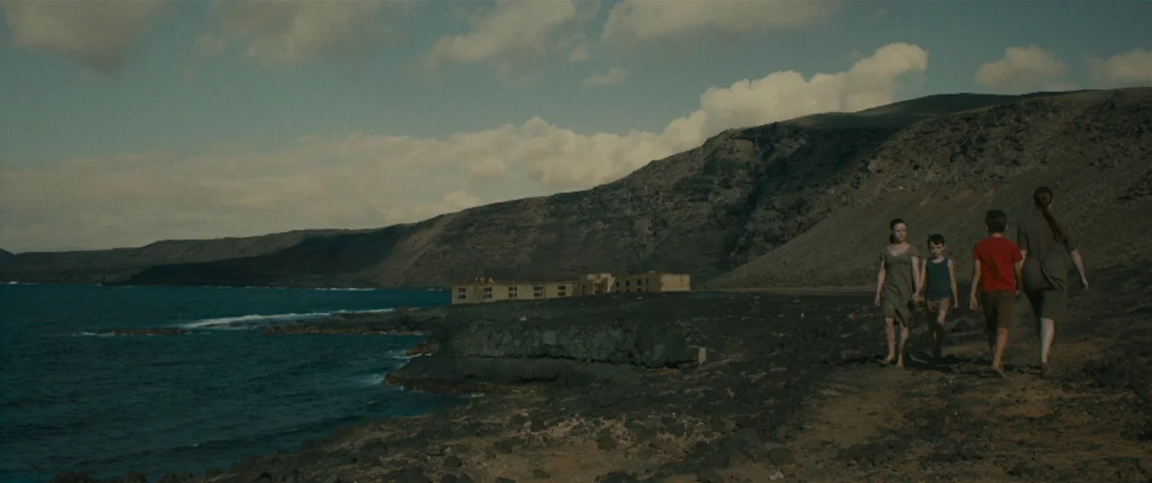 Evolution (2015) movie still: wide — A rugged volcanic coastline stretches alongside a dark blue sea under a cloudy sky. Four individuals walk along…