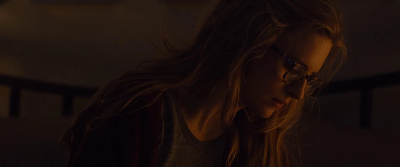 I Origins (2014) movie still: closeup — A dark indoor setting is bathed in low-key, warm ambient illumination. A young woman with glasses looks down…