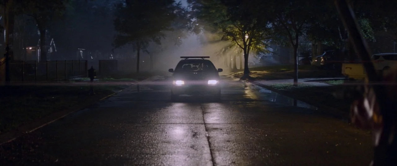 Assassination Nation (2018) movie still: wide — A wet asphalt road in a residential neighborhood is shrouded in heavy night fog. A dark sedan with…