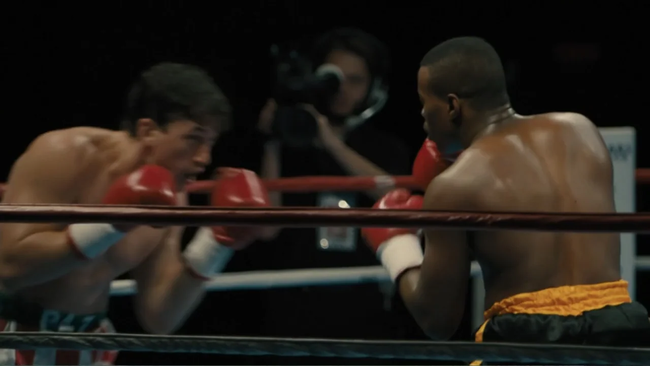 Bleed for This (2016) movie still: medium — A dark boxing ring contains two muscular male boxers engaged in a match. The men wear red gloves and are…