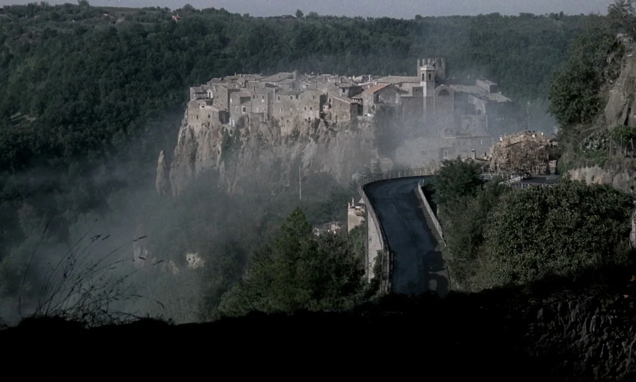 Nostalghia (1983) movie still: establishing — A stone hilltop town sits perched atop a sheer cliff surrounded by dense forest. A curving asphalt…