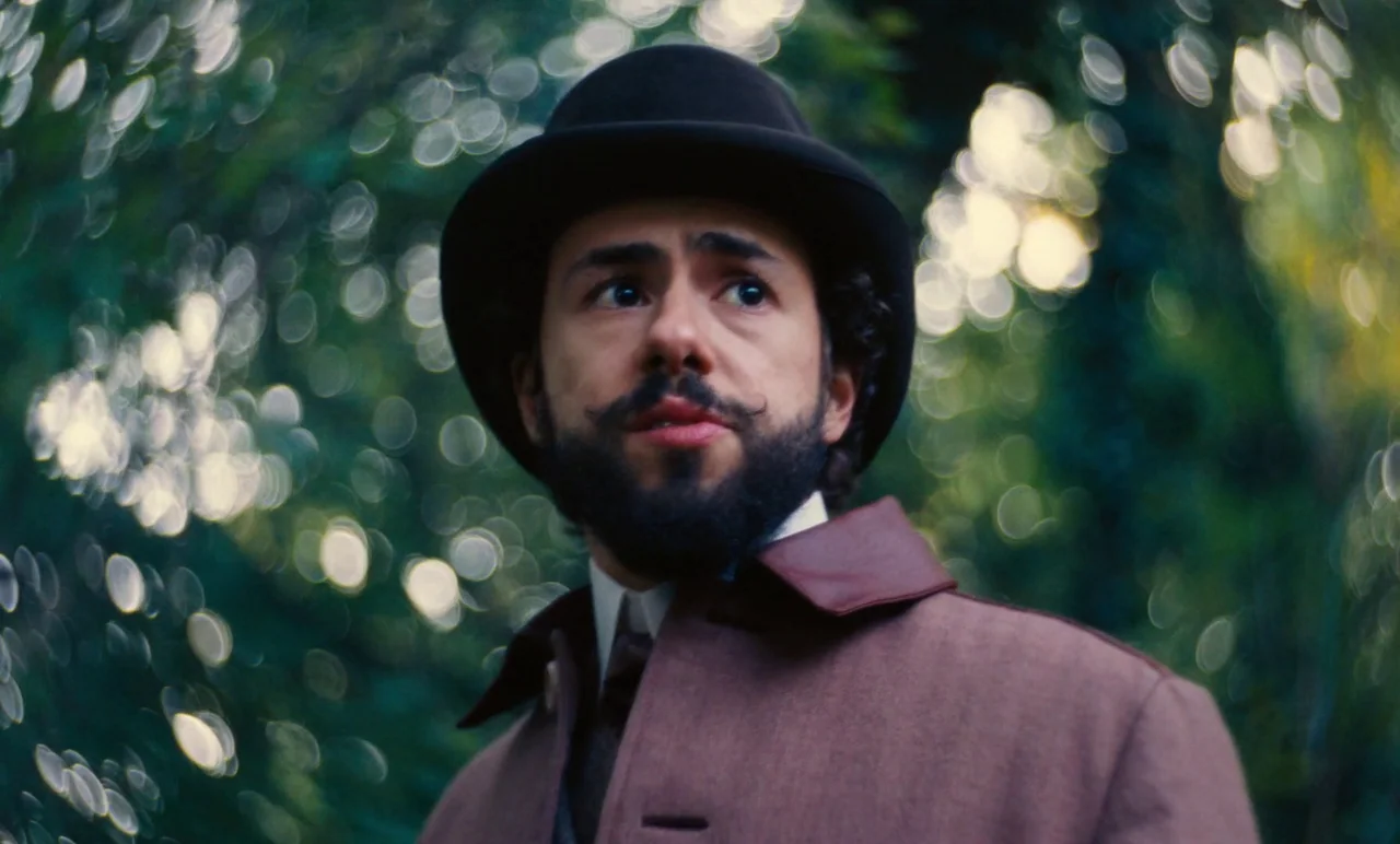 Poor Things (2023) movie still: closeup — A man stands outdoors amidst a blurred backdrop of dense, sun-dappled green leaves. He wears…