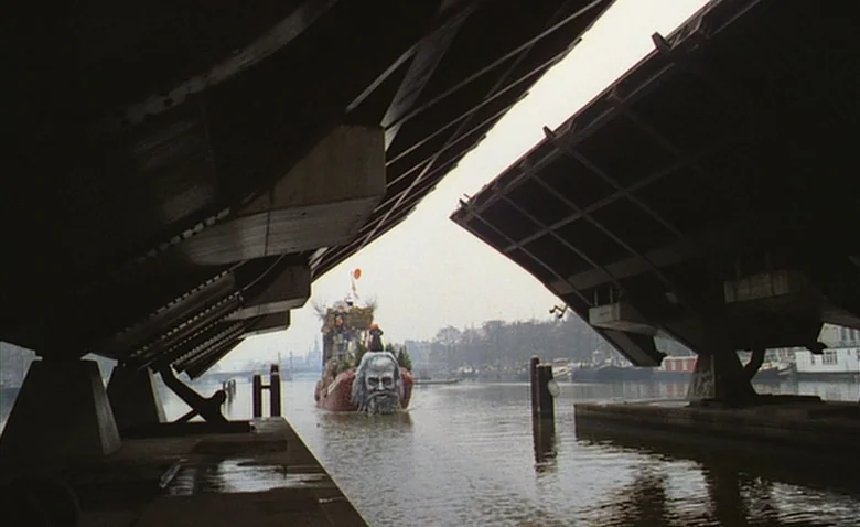 Sweet Movie (1974) movie still: wide — A raised drawbridge forms a dark triangular frame over a river canal. A decorative float adorned with a large…