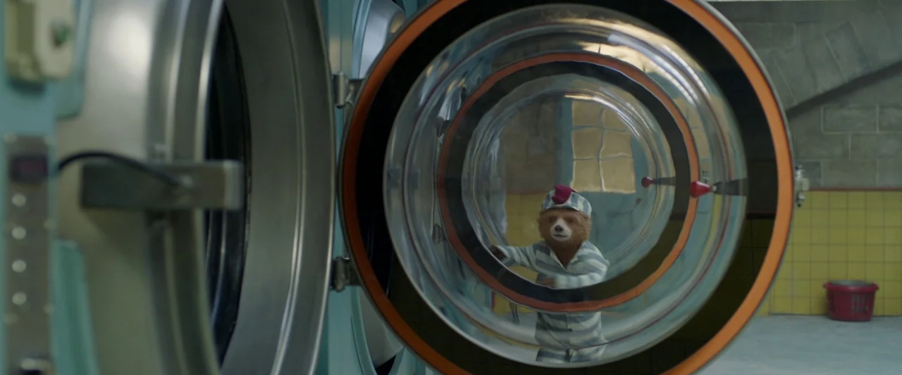 Paddington 2 (2017) movie still: medium — An anthropomorphic bear stands behind the circular glass door of a large washing machine. The background…