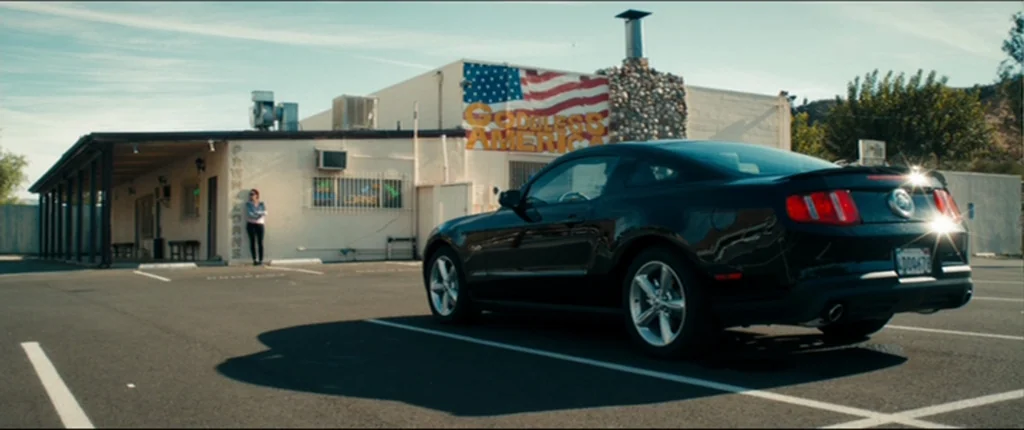 Drive (2011) movie still: wide — A low-slung, single-story building features a large mural depicting an American flag. A woman stands under the…