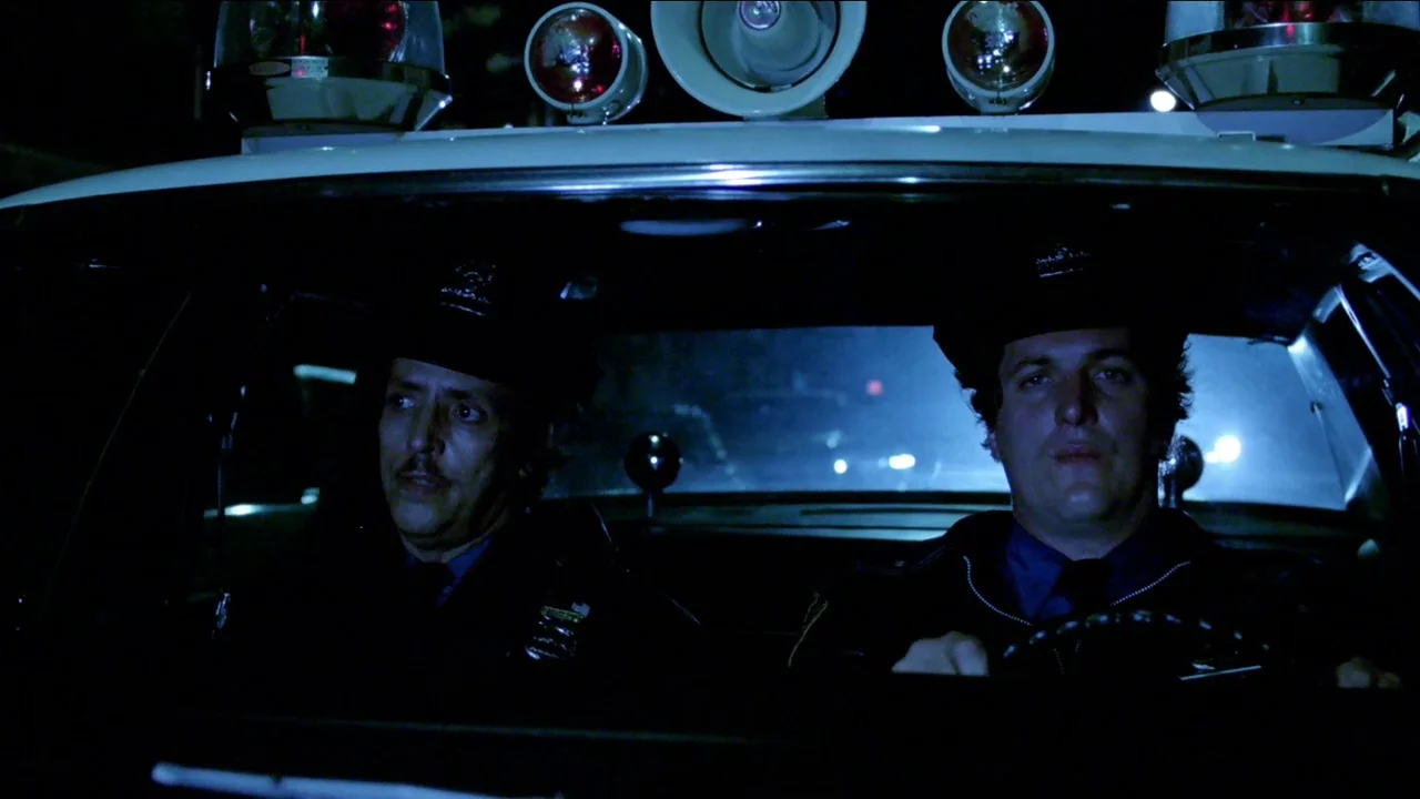 Cruising (1980) movie still: medium — Two uniformed police officers sit inside the front cabin of a patrol vehicle at night. The view looks through…