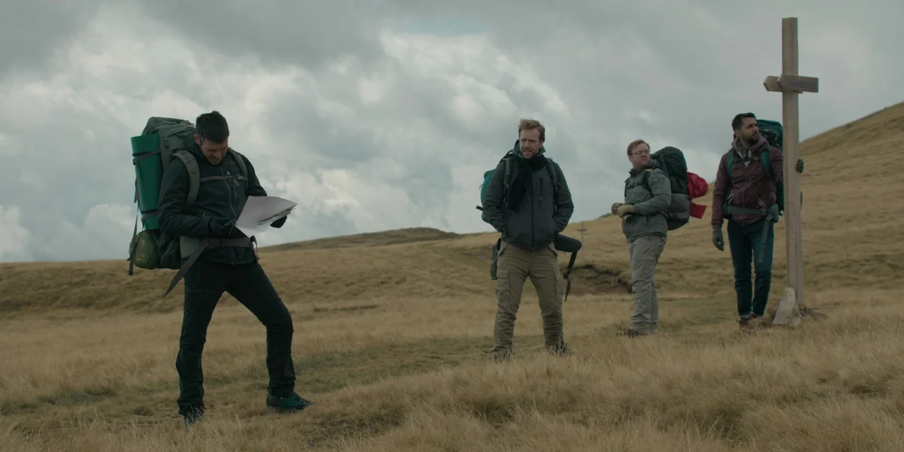 The Ritual (2017) movie still: wide — An open grassy mountain meadow under a heavy, overcast cloudy sky. Four men in hiking gear stand near a wooden…