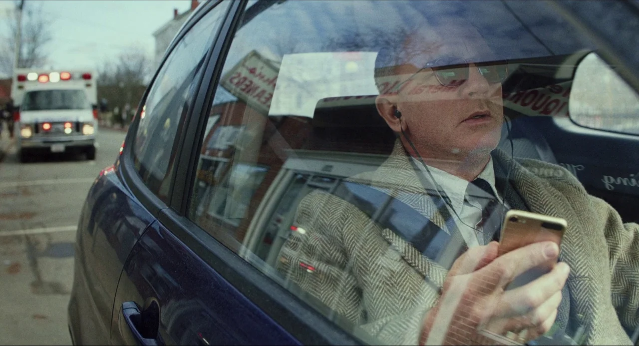 Knives Out (2019) movie still: medium — A man sits in the driver seat of a car looking at his phone. In the background, an ambulance is parked on a…
