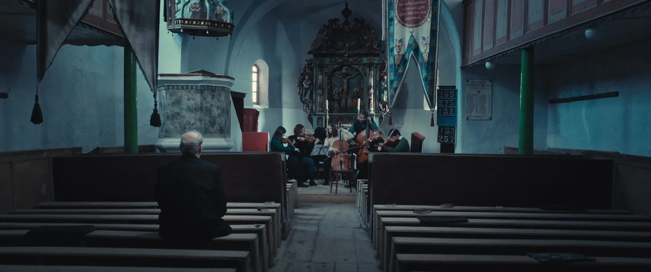 R.M.N. (2022) movie still: wide — A small rustic wooden church features worn pews and an ornate central altar area. A small string ensemble plays…