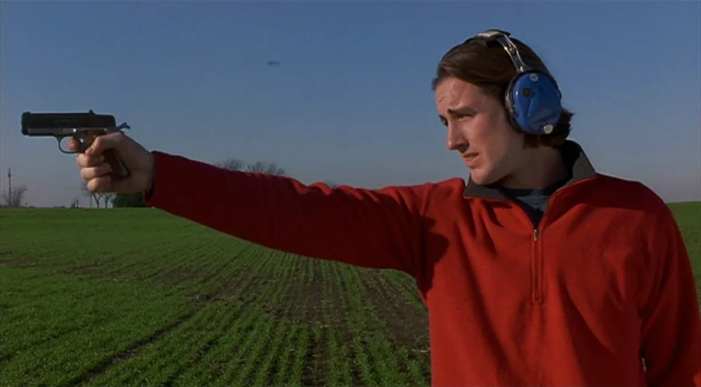 Bottle Rocket (1996) movie still: medium — A man stands in an open agricultural field under a clear blue sky. He aims a handgun toward the left…