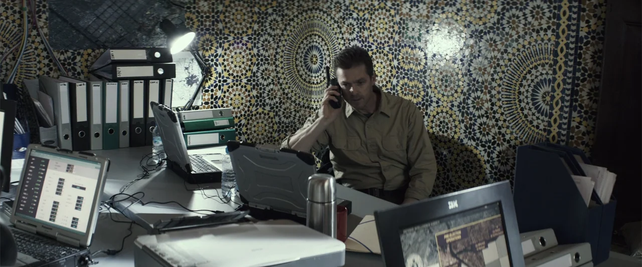 American Sniper (2014) movie still: medium — An office space features intricate geometric tile wall patterns and stacks of file folders. A man in a…