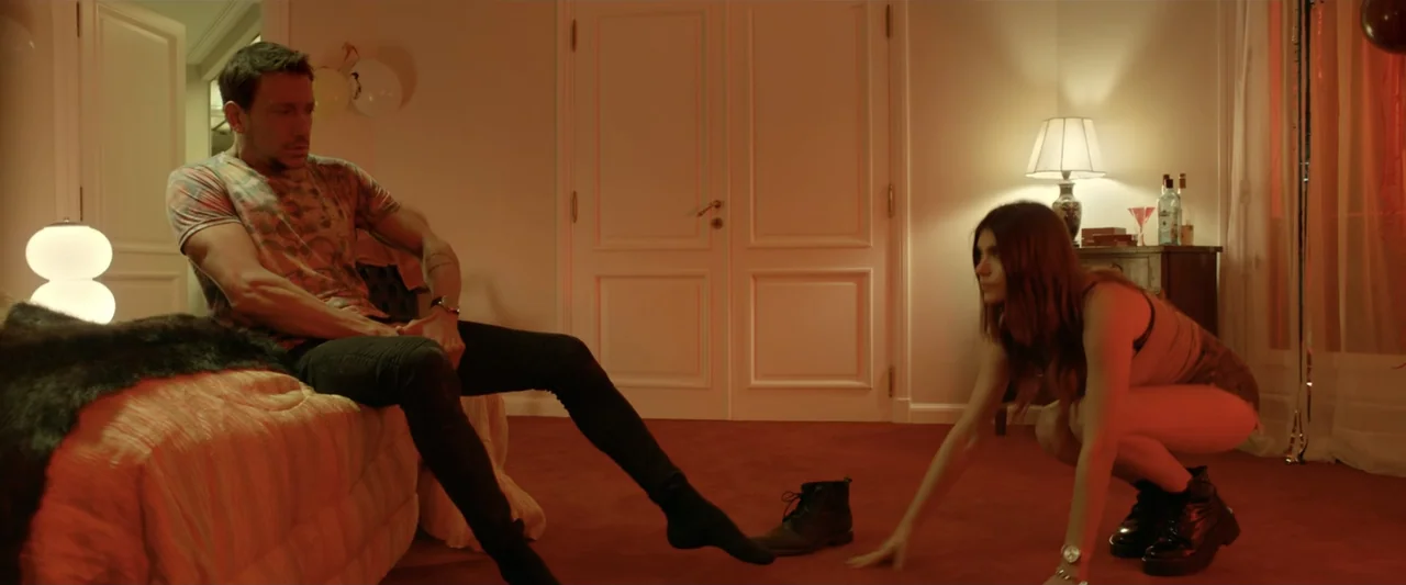 Perdida (2018) movie still: medium — A modern room features red carpeting and white paneled walls with a bedside lamp. A man sits on the edge of the…