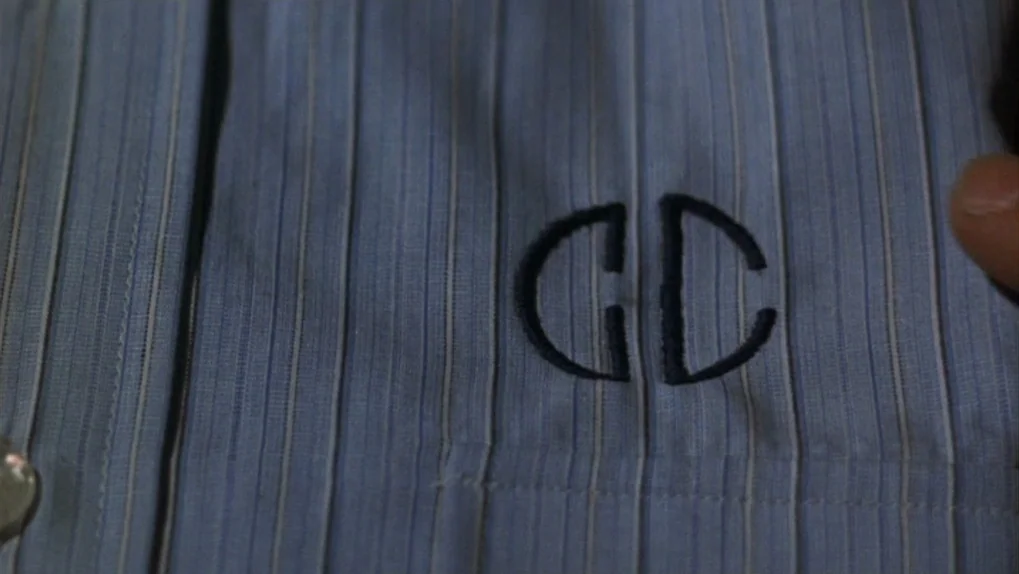 Mean Streets (1973) movie still: closeup — A close-up view of a blue pinstriped dress shirt featuring embroidered monogram initials. A fingertip is…